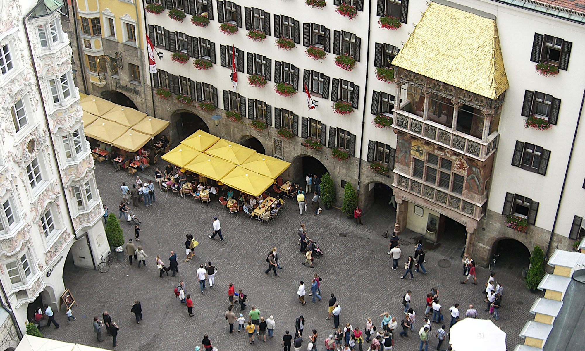 landscape format; view from top of a nearby tower; house facade with lots of windows; in the right half the representative front with balcony and golden roof; on the ground some tables of restaurants and cafes with sunshades, people passing by.