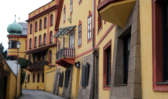 landscape format; tall old building to the right of a road; walls are dark yellow, red frames around the windows, some of the windows are arch-like; dominant colour is yellow;