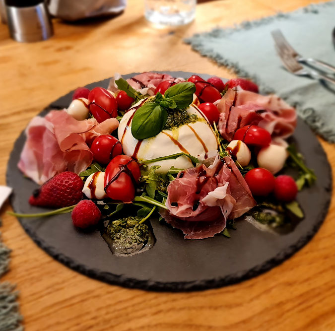 Square format; dominating colour is red; round plate on brown table, on the plate a big ball of burrata, surrounded by raw ham, small tomatoes, strawberries, small balls of burrata, basil, green pesto and balsamic vinegar