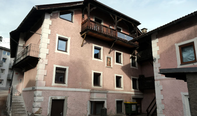 landscape format; big old house, antique pink; 4 storeys, small balconies, one on the front at 4th level, one on the side on 3rd level; in the center of the facade a mural (smaller than a window) of a madonna with child.