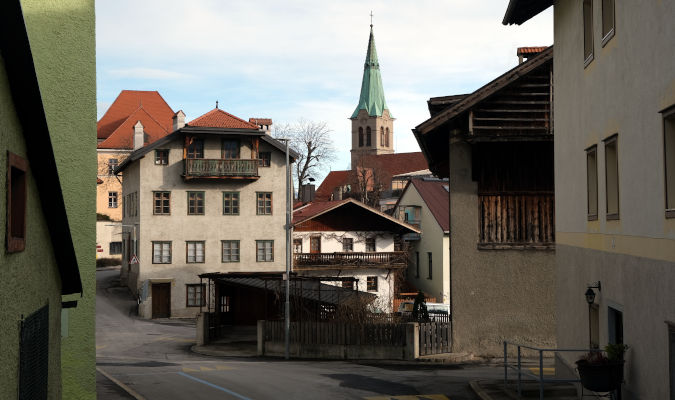 landscape format; road and houses, different sizes, seemingly randomly distributed; brownish grey colours. In the background the green tipped tower of a church