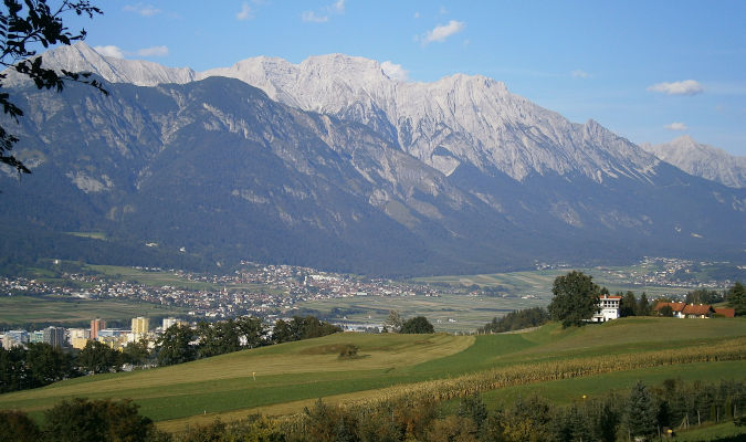 Landscape format; in the foreground a meadow, looking down to a lower level with high rise buildings on the left, regular houses in the middle, the upper half of the picture show mountains. blue sky