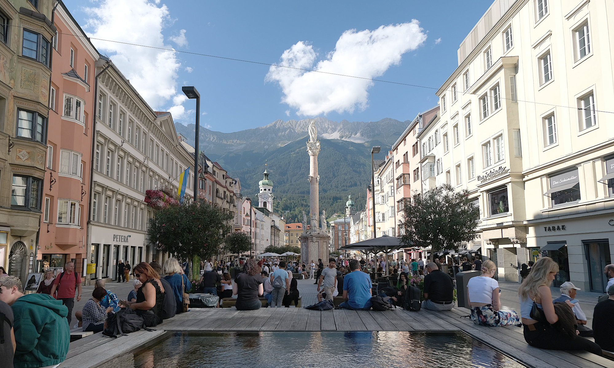 Landscape format; In the foreground a water well, people sitting around it. Left and right rows of houses; in the middle the Anna's column; in the background mountains (Nordkette); it's a warm and sunny day in September 2022