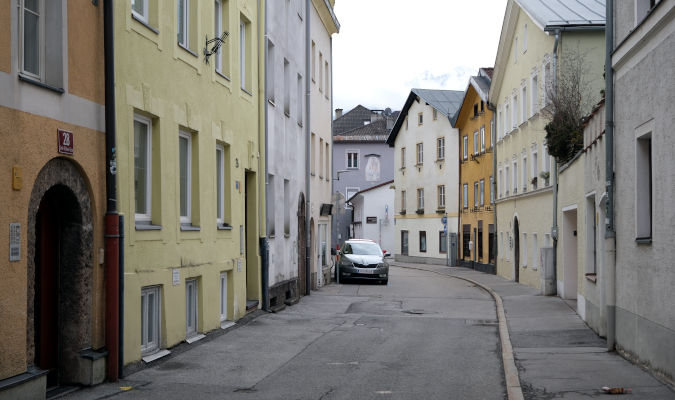 landscape format; small road, empty except for one parking car, Three storey houses in light pastel colours or white or light grey on each side of the road, First house on the left has an archway door, sidewalk on the right. rather boring scene.