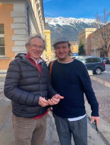 portrait format; two men, 35/60 years old, one with a cap, one with grey hair; dark grey / blue clothes,; background: a road in the city, mountains.