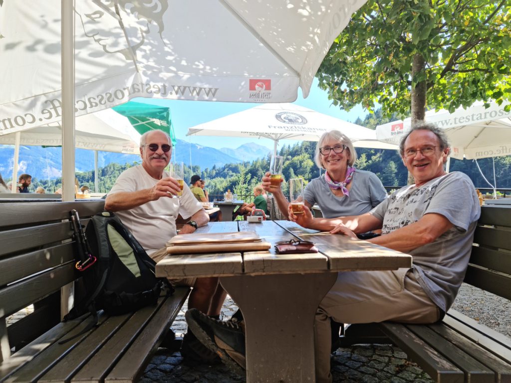 landscape format; table with two benches in open air, two men and one womand look into the camera, holding their beer glasses; sunny day