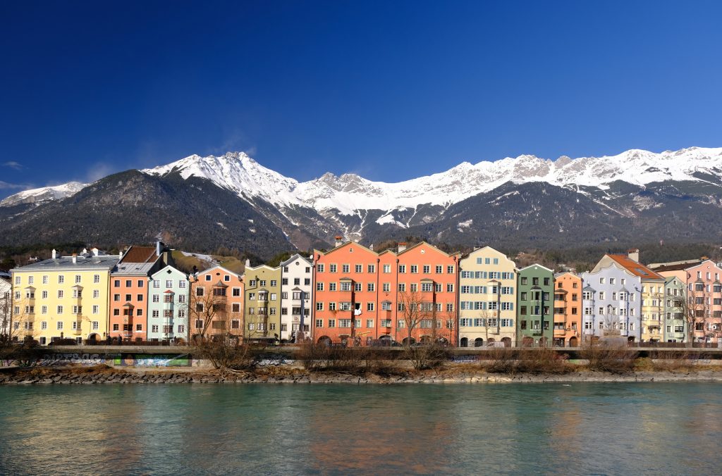Landscape format; in the foreground the river, in the middle a row of coloured houses, behind that the snow capped mountains; sunny