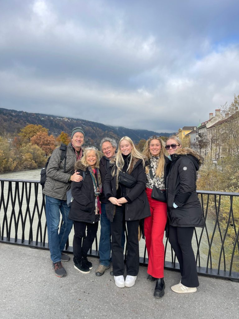 portrait format; 6 people on a bridge, looking into the camera, 3 young women, blonde, in their early 20ies, 2 male and 1 female, in their 50+; winter clothing; cloud covered sky