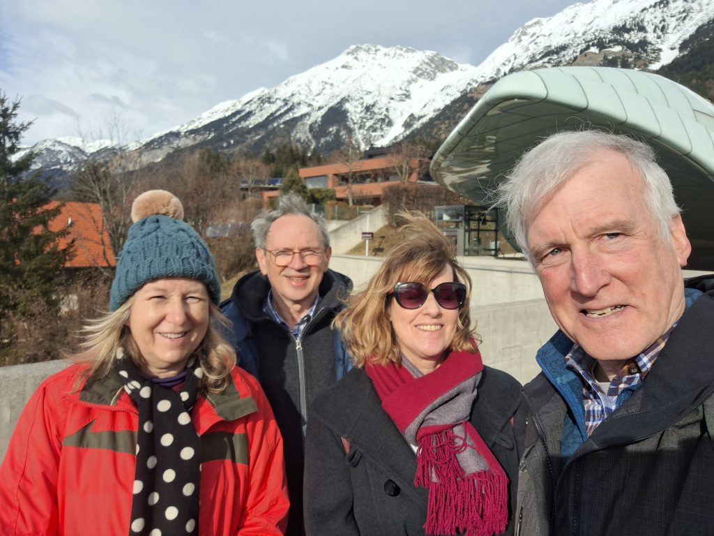 landscape format; selfie with 4 people, from left to right one woman with a woolen hat, red jacket, the rest have dark grey jackets and coats, then one man squinting his eyes, one woman with sunglasses and a red scarf, at the very right a man again. The men have grey hair, the women are dark blonde. In the background snow capped mountains.