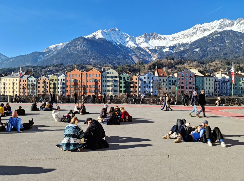 Querformat; großer Platz, Fußgängerzone; sonniger Frühlingstag; viele junge Leute sitzen auf dem Asphalt; in der Mitte (vertikal) des Bildes die bunte Häuserzeile; im Hintergrund schneebedeckte Berge