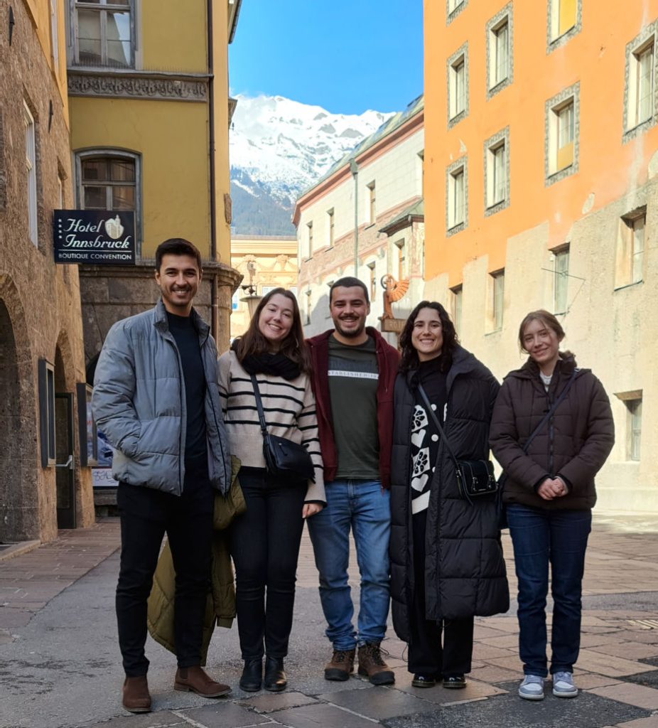 square format; five young people (two men, three women), full body photo, smiling into the camera. In the background old houses, yellow and orange; further back the snow capped mountains