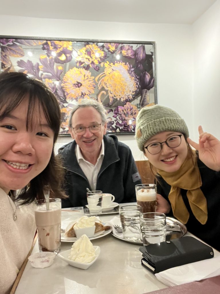 portrait format, selfie; 3 people at a table in a Cafe; left a young woman, middle an older man. right a young woman again. On the table various coffees, in the background a colourful painting of flowers