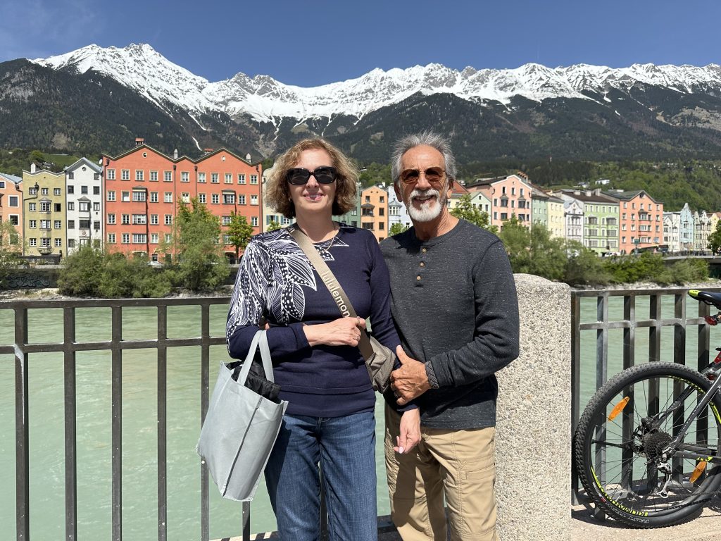 landscape format; a woman (ca 50 yrs) and a man (ca 65 yrs), both wearing trousers and shirts, sunglasses, smile into the camera. behind them the river, a row of coloured houses and the mountains