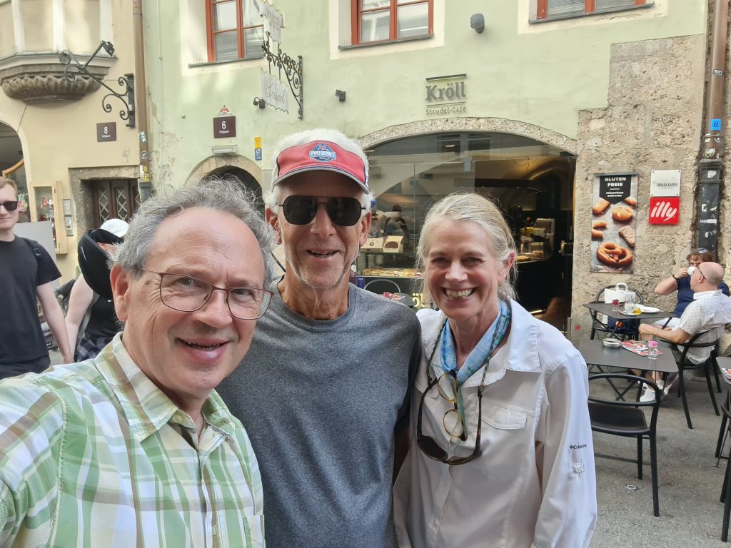 landscape format; 3 people on a selfie, male, male, female, ca 60-70 years, first with green shirt, glasses, grey hair, 2nd grey shirt, sunglasses, red visor, 3rd light grey jacket, grey hair; all smiling of course, in the background street scene, green house, tables of a cafe