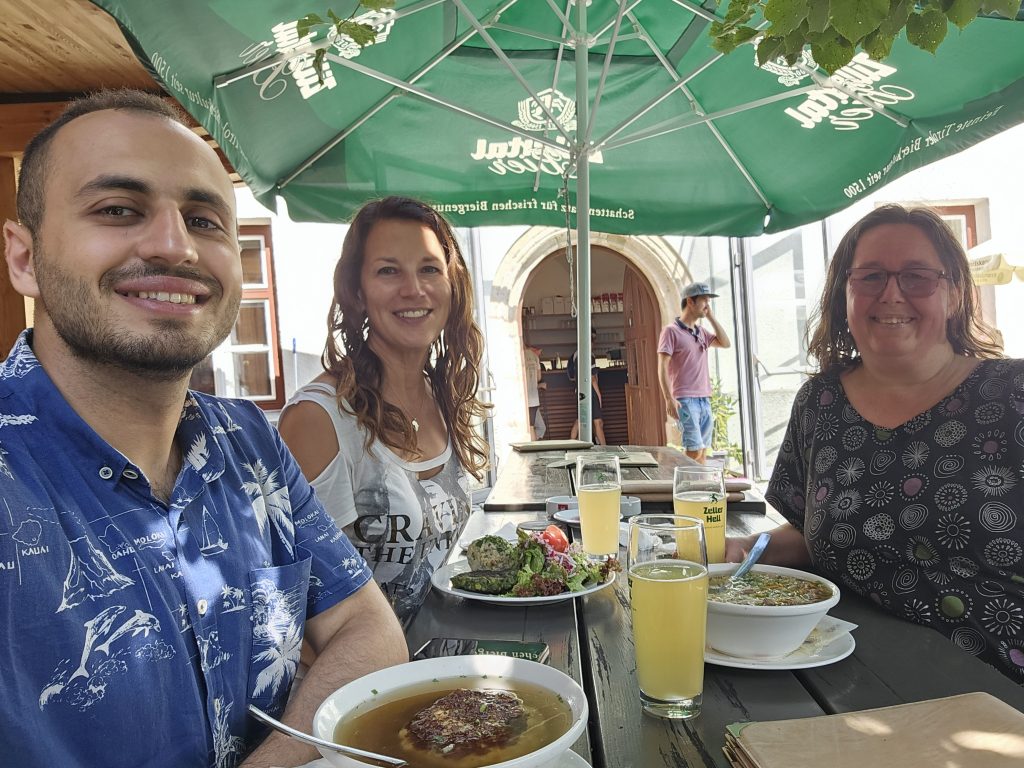 landscape format; selfie of 3 people at a table, open air, with their food; man with blue shirt and a beard, woman with white t-shirt, decorated, long hair, woman with dark grey dress, looking glasses